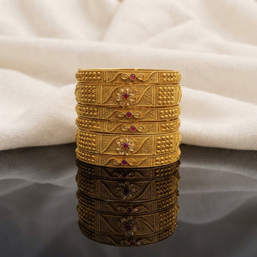 Gold Antique Polki Leaf Vine Floral Ruby Wide Bangles Set of 6 in 22K antique gold-tone plating on dark grey mirror surface with ivory cream fabric backdrop showing six wide rigid bangles stacked together with 3-section band featuring dense gold seed bead granulation panels on sides and diagonal twisted vine filigree with oval polki leaf stones and 3D daisy flower with ruby red cabochon centre with mirror reflection below