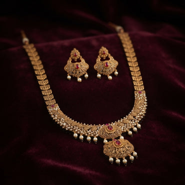 Gold Antique Ruby CZ Filigree Leaf Chain Necklace and Chandbali Earrings Set flat lay on grey mirror surface with dark grey satin backdrop showing 2-piece set with long graduated filigree leaf-link chain necklace with scattered ruby CZ accents and double-tiered Mughal pendant with pearl fringe drops and matching Chandbali earrings with mirror reflection