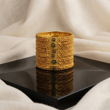 Gold Antique Seed Bead Diamond Frame Ruby Emerald Bangles Set of 4 in 22K antique gold-tone plating on dark black diamond-shaped mirror tray with ivory cream fabric backdrop showing four wide rigid bangles stacked together with dense multi-row gold seed bead granulation texture and diamond-shape raised gold frame centre panels with 4-petal flower motifs in alternating emerald green square and ruby red round cabochon centres with mirror reflection below