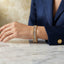 Gold Baguette CZ Channel-Set Double Ball-Edge Wide Tennis Cuff Bracelet worn on model wrist resting on white marble table with beige wall backdrop and navy blue blazer showing full baguette CZ center channel row with double gold ball bead borders on wide rigid cuff