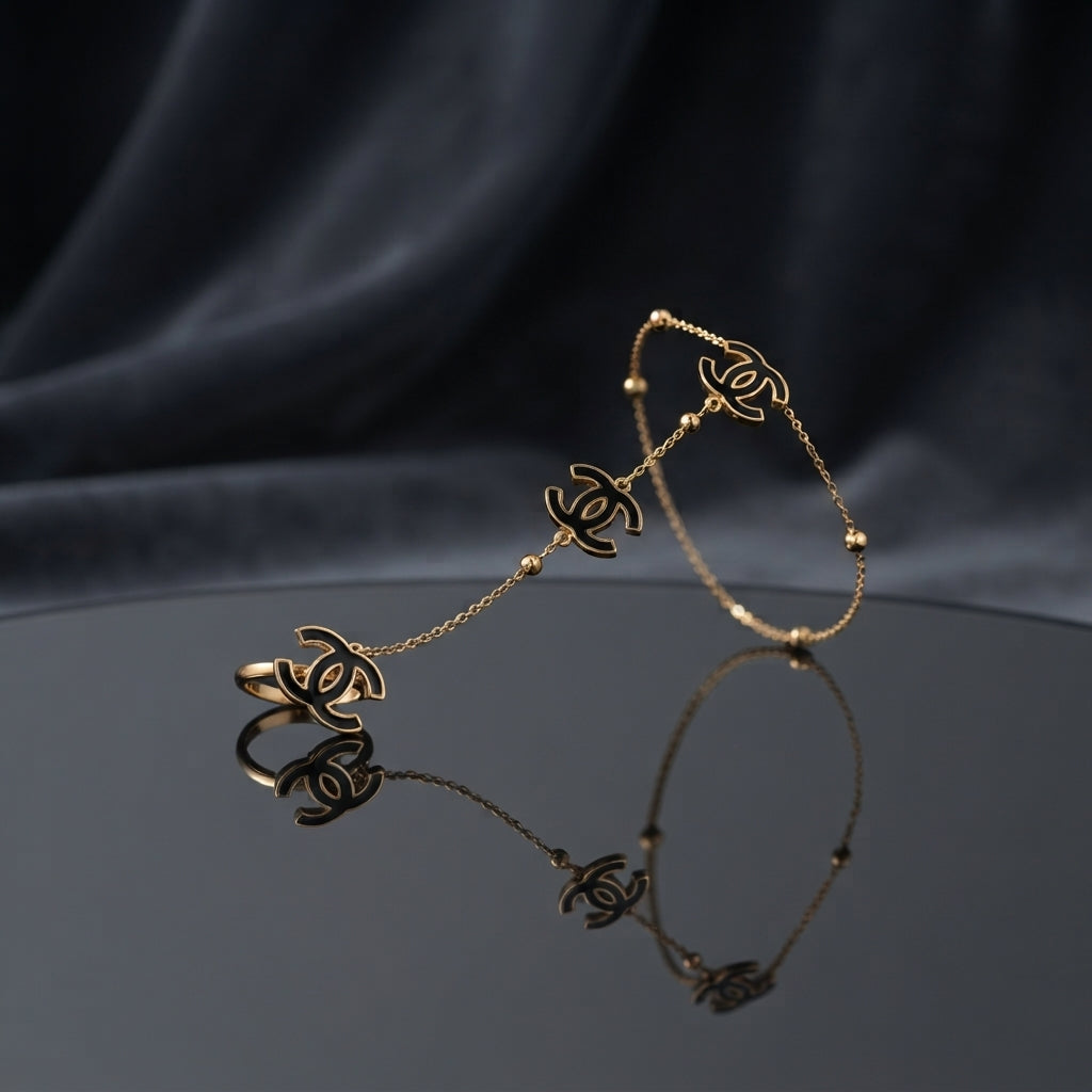 Gold Black Enamel Double-C Logo Station Bracelet and Ring Set in 18K gold-tone PVD stainless steel on dark grey mirror surface with black silk backdrop showing slim satellite station chain bracelet with 3 graduating double-C interlocking logo charms in black enamel and matching adjustable open ring with large double-C logo charm with mirror reflection below