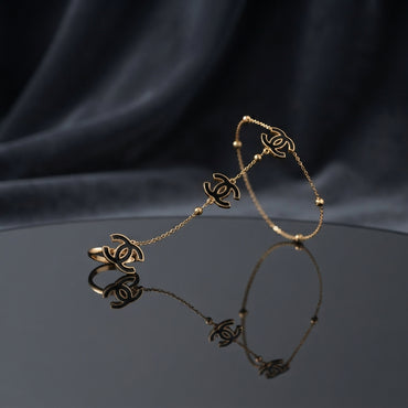 Gold Black Enamel Double-C Logo Station Bracelet and Ring Set in 18K gold-tone PVD stainless steel on dark grey mirror surface with black silk backdrop showing slim satellite station chain bracelet with 3 graduating double-C interlocking logo charms in black enamel and matching adjustable open ring with large double-C logo charm with mirror reflection below