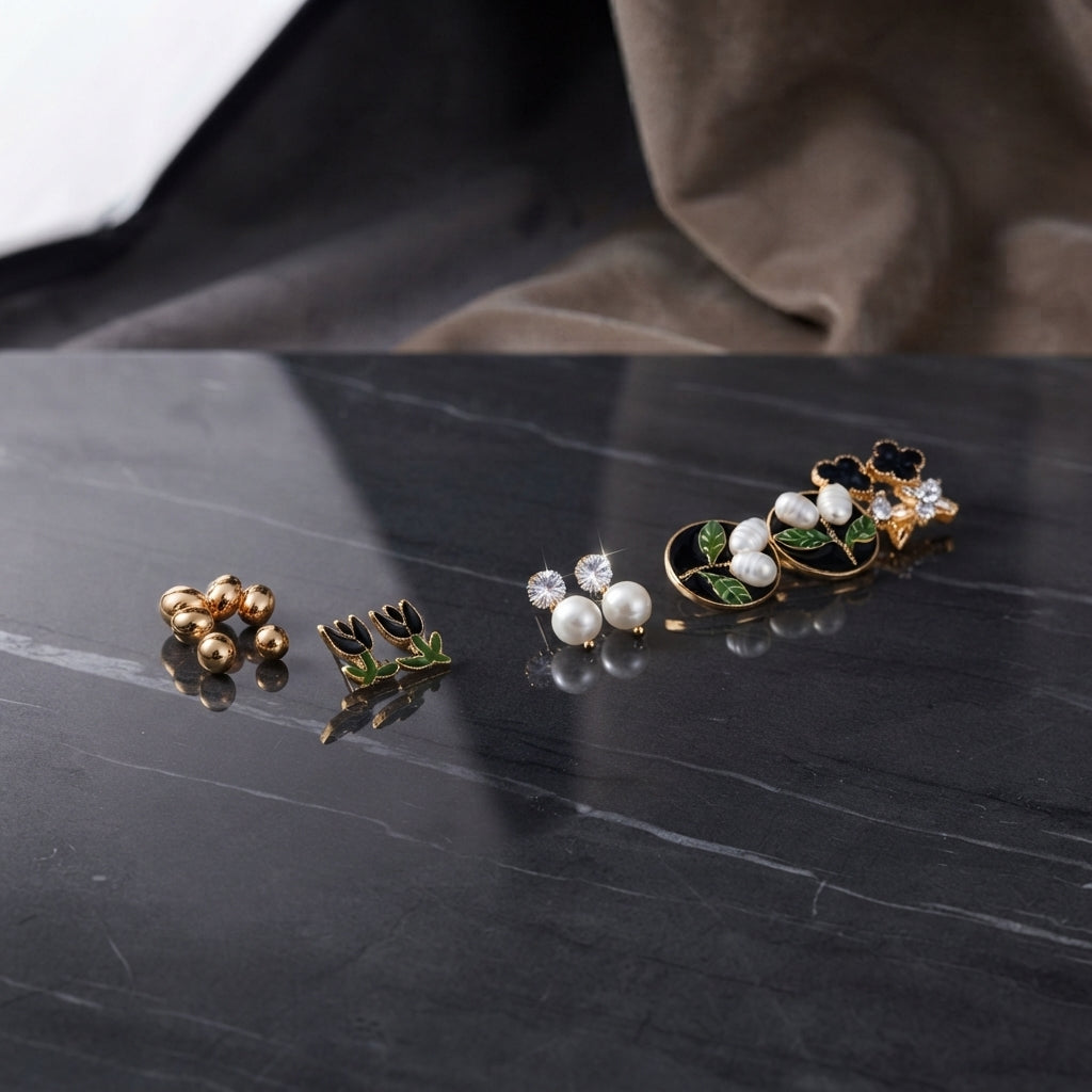 Gold Black Floral Enamel Pearl CZ 4-Pair Stud Earring Set displayed in open clear acrylic gift box on black marble surface with grey fabric backdrop showing all four pairs arranged on white velvet insert with mirror reflection below
