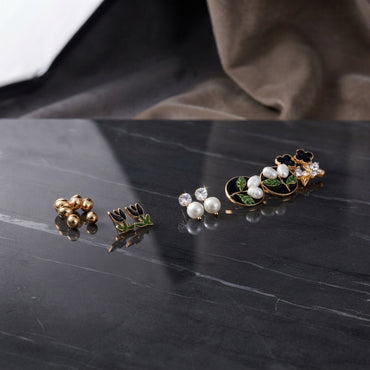 Gold Black Floral Enamel Pearl CZ 4-Pair Stud Earring Set displayed in open clear acrylic gift box on black marble surface with grey fabric backdrop showing all four pairs arranged on white velvet insert with mirror reflection below