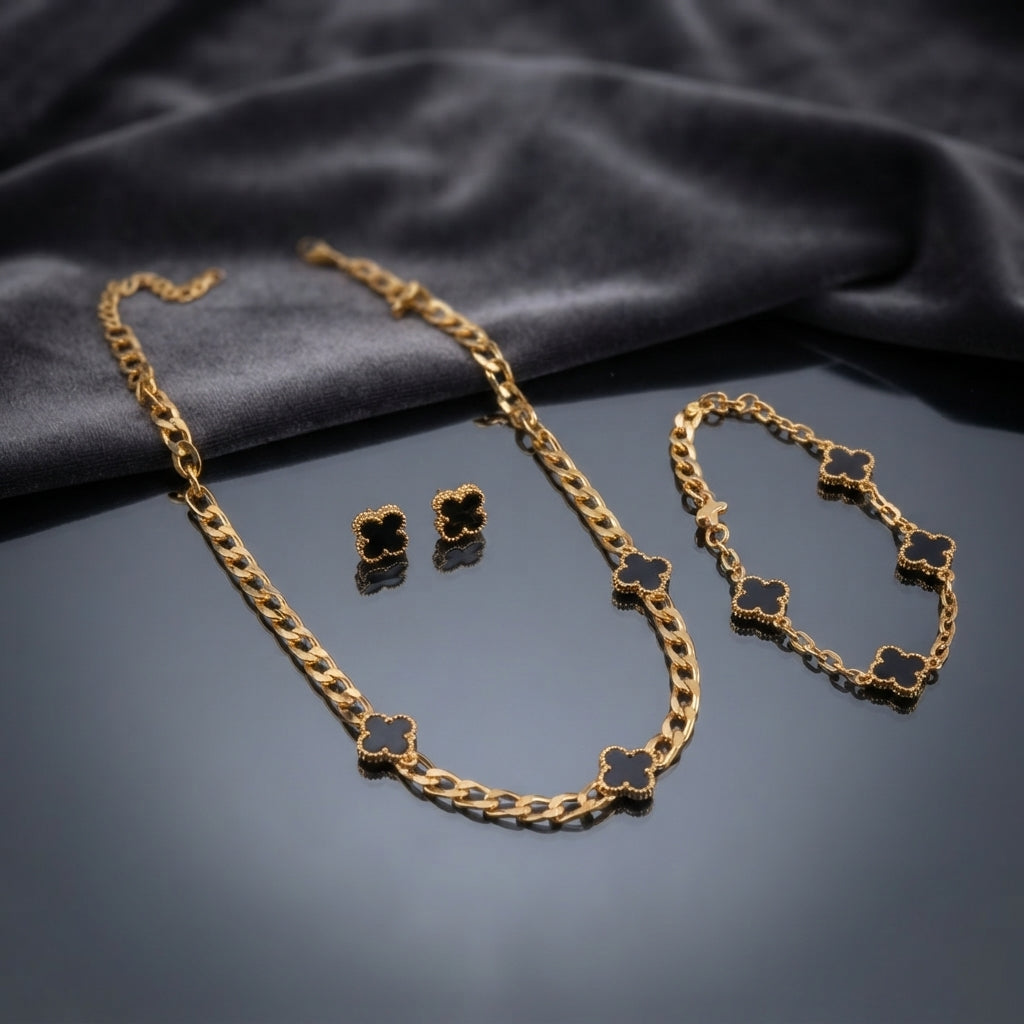 Gold Black Onyx Milgrain 4-Leaf Clover Figaro Chain Necklace Bracelet Earrings Set — 3-piece matching set of 22K gold-tone Figaro chain necklace with 5 solid black onyx milgrain clover charms, matching bracelet with 4 clover charms, and black onyx milgrain clover stud earrings, luxury flat-lay on mirror surface with dark charcoal silk backdrop