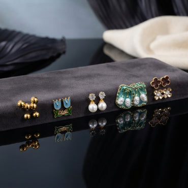 Gold Blue Tulip Floral Enamel Pearl CZ 4-Pair Stud Earring Set flat lay on grey velvet tray with dark blue and cream fabric backdrop showing gold double ball studs blue sky tulip enamel studs with green leaf white CZ pearl drop studs and large teal green enamel square stud with baroque faux pearl drops green leaf brown clover and CZ snowflake cluster with mirror reflections below