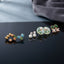 Gold Blue Tulip Mint Floral Pearl Green Clover 4-Pair Stud Earring Set flat lay on dark black mirror surface with grey velvet backdrop showing gold pebble arc studs blue tulip enamel studs with green leaf white CZ pearl drop studs large mint blue enamel disc stud with pearl drops and green clover blush pink CZ flower cluster stud with mirror reflections below