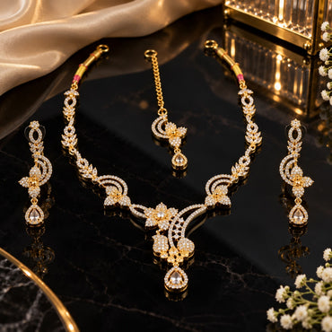 Gold Champagne CZ Floral Vine Necklace Maang Tikka and Linear Drop Earrings Bridal Set — 22K gold-tone 3-piece bridal set with open floral vine necklace with 4-petal CZ flower clusters infinity scroll connectors and champagne pear CZ V-drop matching maang tikka with champagne pear CZ halo drop and 3-tier infinity scroll floral drop earrings displayed on dark mirror surface with black satin backdrop