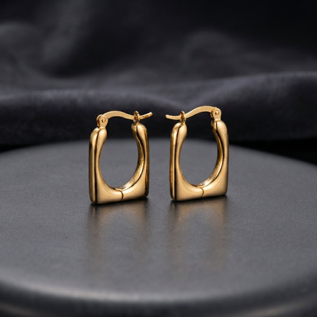 Gold Chunky Square Cushion Hoop Earrings pair standing upright on dark charcoal ceramic plate with dark grey satin backdrop showing 18K gold-tone high-polish mirror finish graduated tubular square hoops with rounded inner corners and hinged click closure in warm editorial lighting