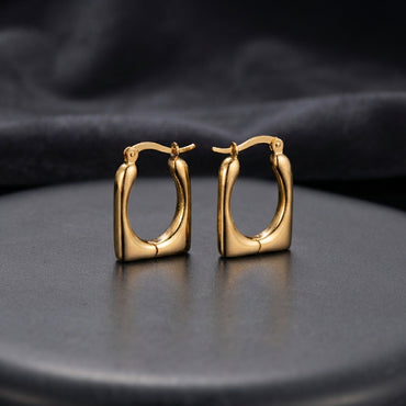 Gold Chunky Square Cushion Hoop Earrings pair standing upright on dark charcoal ceramic plate with dark grey satin backdrop showing 18K gold-tone high-polish mirror finish graduated tubular square hoops with rounded inner corners and hinged click closure in warm editorial lighting