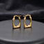 Gold Chunky Square Cushion Hoop Earrings pair standing upright on dark charcoal ceramic plate with dark grey satin backdrop showing 18K gold-tone high-polish mirror finish graduated tubular square hoops with rounded inner corners and hinged click closure in warm editorial lighting