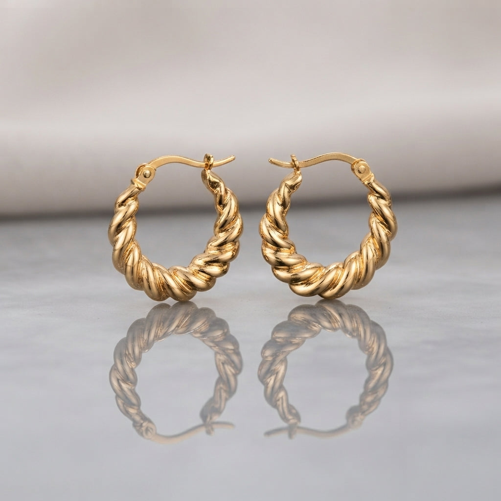 Gold PVD Stainless Steel Chunky Twisted Rope Croissant Hoop Earrings pair standing side by side on light grey mirror surface with soft warm cream ivory fabric backdrop showing 18K gold PVD coated stainless steel hoops with thick twisted rope croissant design alternating satin-brushed valleys and mirror-polish ridges graduated width thicker at bottom with French lever closure and soft mirror reflection below