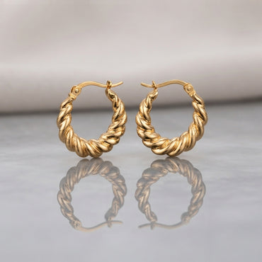Gold PVD Stainless Steel Chunky Twisted Rope Croissant Hoop Earrings pair standing side by side on light grey mirror surface with soft warm cream ivory fabric backdrop showing 18K gold PVD coated stainless steel hoops with thick twisted rope croissant design alternating satin-brushed valleys and mirror-polish ridges graduated width thicker at bottom with French lever closure and soft mirror reflection below