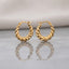 Gold PVD Stainless Steel Chunky Twisted Rope Croissant Hoop Earrings pair standing side by side on light grey mirror surface with soft warm cream ivory fabric backdrop showing 18K gold PVD coated stainless steel hoops with thick twisted rope croissant design alternating satin-brushed valleys and mirror-polish ridges graduated width thicker at bottom with French lever closure and soft mirror reflection below