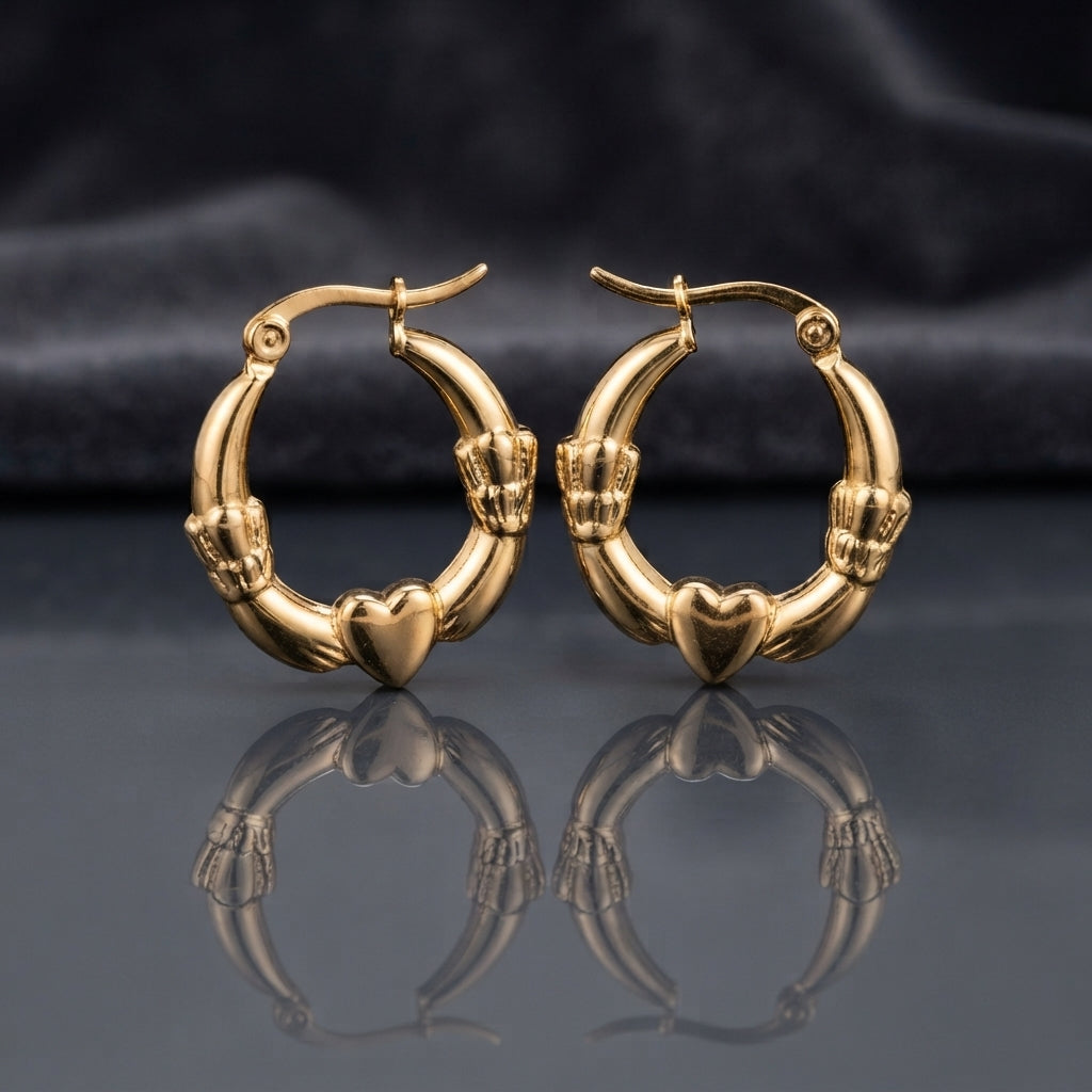 Gold Claddagh Heart and Hands Hoop Earrings pair standing side by side on dark mirror surface with charcoal slate satin backdrop showing 22K gold-tone mirror-polish hoops with smooth puffy 3D heart at base held by two detailed sculpted hands with engraved finger detail on both sides in classic Claddagh arrangement with sharp mirror reflection below