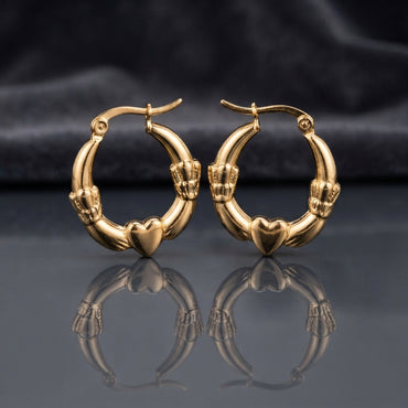 Gold Claddagh Heart and Hands Hoop Earrings pair standing side by side on dark mirror surface with charcoal slate satin backdrop showing 22K gold-tone mirror-polish hoops with smooth puffy 3D heart at base held by two detailed sculpted hands with engraved finger detail on both sides in classic Claddagh arrangement with sharp mirror reflection below