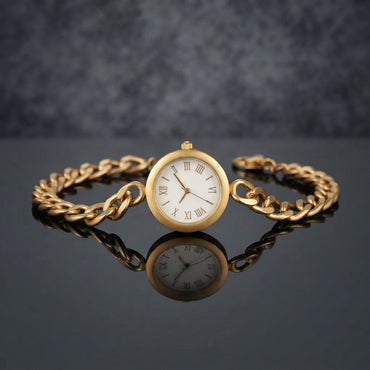 Gold Cuban Link Chain Bracelet Watch in 18K gold-tone PVD stainless steel on dark grey mirror surface with grey bokeh backdrop showing bold Cuban curb link chain bracelet with smooth round polished gold-tone watch case and decorative white Roman numeral watch-face dial sticker with gold-tone hands and mirror reflection below