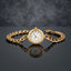 Gold Cuban Link Chain Bracelet Watch in 18K gold-tone PVD stainless steel on dark grey mirror surface with grey bokeh backdrop showing bold Cuban curb link chain bracelet with smooth round polished gold-tone watch case and decorative white Roman numeral watch-face dial sticker with gold-tone hands and mirror reflection below