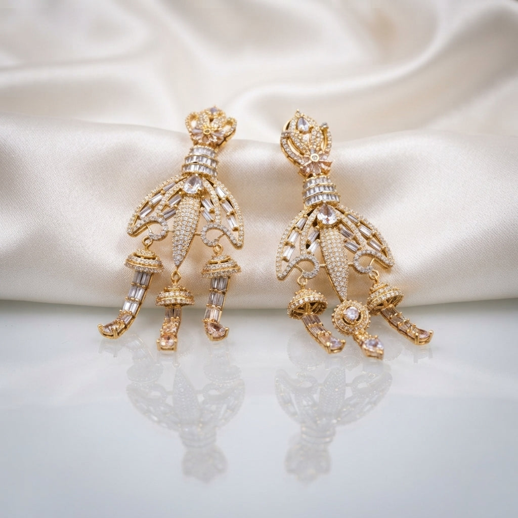 Gold CZ Champagne Baguette Crown and Chandelier Fan Jhumka Drop Earrings on white ivory satin backdrop showing 22K warm gold-tone grand chandelier jhumka pair with ornate lotus crown top baguette CZ ribbed column open-work fan chandelier body with champagne CZ arch arms 3 tiered mini jhumka bells and champagne morganite pear CZ drops