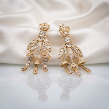 Gold CZ Champagne Baguette Crown and Chandelier Fan Jhumka Drop Earrings on white ivory satin backdrop showing 22K warm gold-tone grand chandelier jhumka pair with ornate lotus crown top baguette CZ ribbed column open-work fan chandelier body with champagne CZ arch arms 3 tiered mini jhumka bells and champagne morganite pear CZ drops