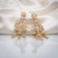 Gold CZ Champagne Baguette Crown and Chandelier Fan Jhumka Drop Earrings on white ivory satin backdrop showing 22K warm gold-tone grand chandelier jhumka pair with ornate lotus crown top baguette CZ ribbed column open-work fan chandelier body with champagne CZ arch arms 3 tiered mini jhumka bells and champagne morganite pear CZ drops