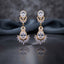 Gold CZ Double Teardrop Marquise Fan Chandelier Earrings in Jet Black pair on dark mirror surface with grey silk backdrop showing 18K gold-tone small black pear CZ stud with white CZ pave halo infinity connector and large black pear CZ drop with white CZ pave halo and marquise CZ fan crown with mirror reflection below