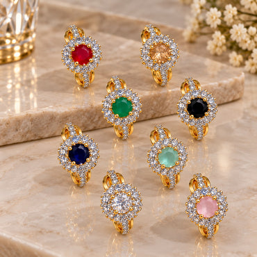 Gold CZ Halo Cocktail Ring with oval coloured CZ centre stone in double white CZ halo cluster and triple split-shank pave band shown in all 9 color variants Ruby Red Champagne Emerald Green Jet Black Navy Blue Mint Green Crystal Clear Baby Pink and Teal arranged on warm marble surface with white floral backdrop in 18K gold-tone plated finish