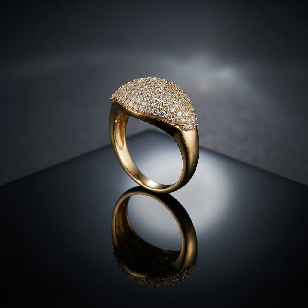 Gold CZ Micro-Pavé Cushion Dome Signet Cocktail Ring — 22K gold-tone modern luxury signet ring with large cushion oval dome fully covered in dense round white CZ micro-pavé, smooth seamless transition to high-polish tapered gold shank, on dark grey mirror surface with luxury editorial backdrop