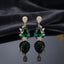 Gold CZ Pear Halo and Marquise Floral Vine Oval Crystal Drop Earrings in Emerald Green on dark charcoal slate satin with dark mirror surface showing 22K gold-tone pair with pear white CZ halo stud deep vivid emerald green faceted oval crystal pendant marquise CZ floral vine sprigs on both sides and 5-petal marquise CZ daisy flower base