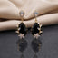 Gold CZ Pear Halo and Marquise Floral Vine Oval Crystal Drop Earrings in Jet Black on warm cream ivory satin with dark slate mirror showing 22K gold-tone pair with pear white CZ halo stud deep jet black onyx faceted oval crystal pendant marquise CZ floral vine sprigs on both sides and 5-petal marquise CZ daisy flower base