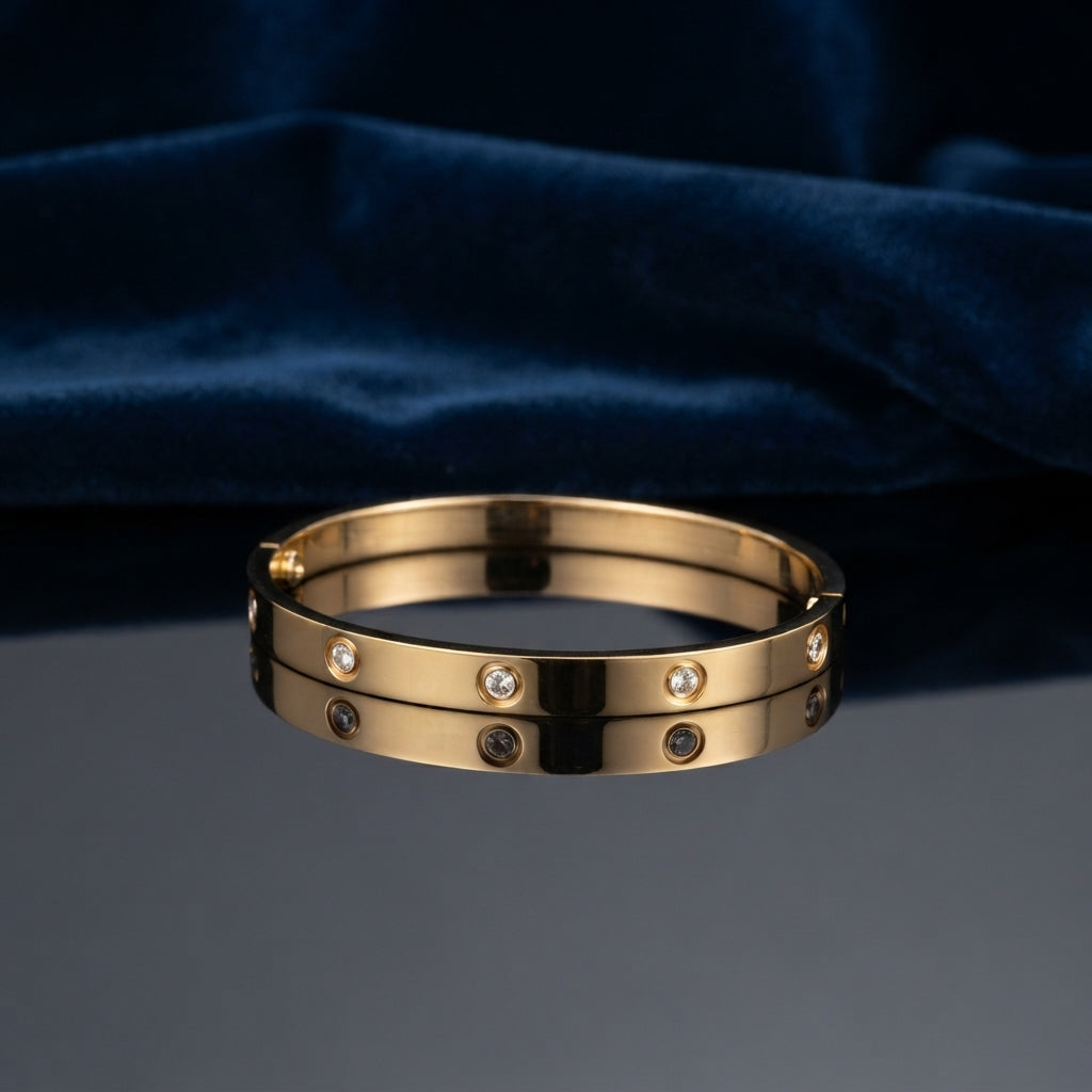 Gold CZ Studded Screw Motif Hinged Bangle Bracelet worn on right wrist of female model in navy blue blazer and white top with hands clasped on dark wood table showing 18K warm yellow gold-tone hinged bangle with alternating screw motif bezels and flush bezel-set round white CZ stones in luxury boutique setting
