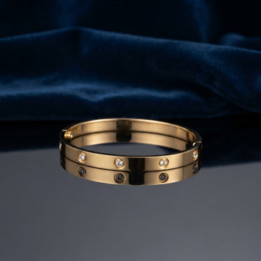 Gold CZ Studded Screw Motif Hinged Bangle Bracelet worn on right wrist of female model in navy blue blazer and white top with hands clasped on dark wood table showing 18K warm yellow gold-tone hinged bangle with alternating screw motif bezels and flush bezel-set round white CZ stones in luxury boutique setting