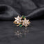 Gold Double Flower Cluster CZ Cocktail Ring in deep vivid emerald green CZ accent variant — luxury flat-lay on mirror surface with charcoal silk backdrop