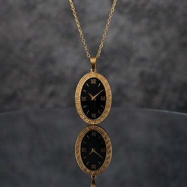 Gold Elongated Oval Greek Key Black Dial Watch-Face Pendant Necklace in 18K gold-tone PVD stainless steel on dark grey mirror surface with grey textured backdrop showing tall portrait oval pendant with continuous Greek key meander engraved border and deep black decorative Roman numeral watch-face dial sticker with gold-tone hands on slim gold cable chain with mirror reflection below