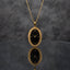 Gold Elongated Oval Greek Key Black Dial Watch-Face Pendant Necklace in 18K gold-tone PVD stainless steel on dark grey mirror surface with grey textured backdrop showing tall portrait oval pendant with continuous Greek key meander engraved border and deep black decorative Roman numeral watch-face dial sticker with gold-tone hands on slim gold cable chain with mirror reflection below