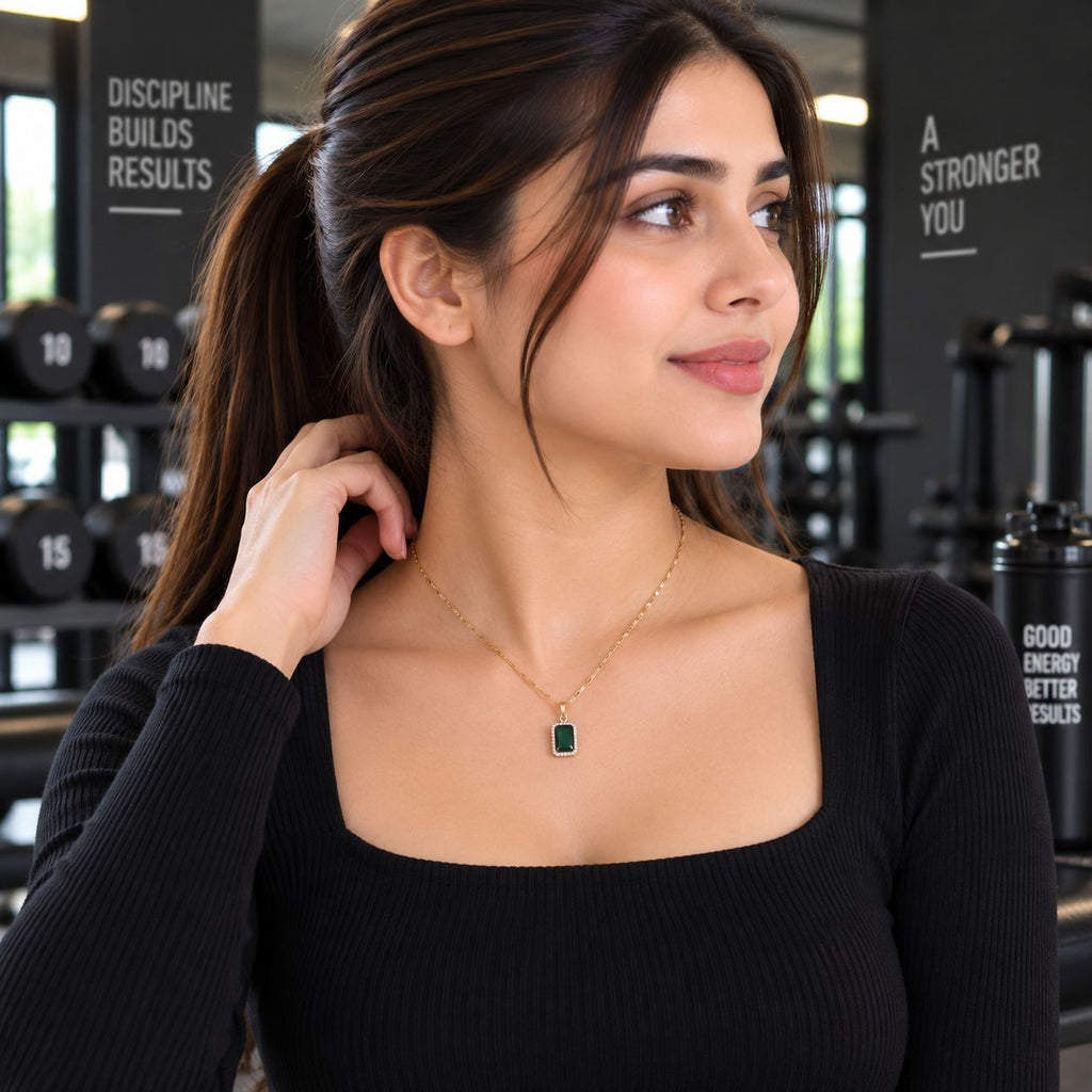 Gold Emerald Rectangle CZ Halo Pendant Necklace worn by model in black long-sleeve top at gym setting with dumbbell rack backdrop