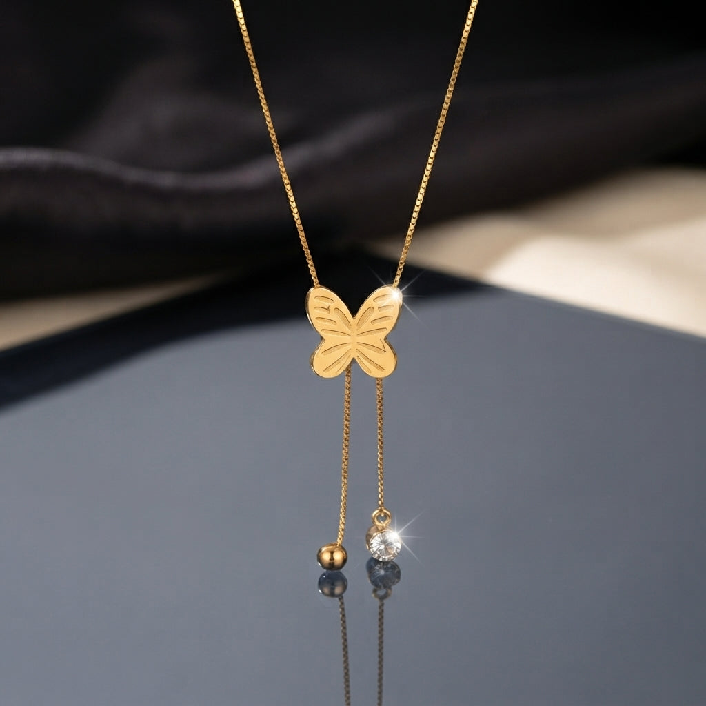 Gold Engraved Butterfly Tassel CZ Ball Drop Lariat Necklace flat lay on dark grey mirror surface with dark silk and beige fabric backdrop showing engraved gold butterfly pendant with intricate wing line details and double tassel drop chains with gold ball charm and round bezel CZ solitaire drop with mirror reflection below