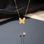 Gold Engraved Butterfly Tassel CZ Ball Drop Lariat Necklace flat lay on dark grey mirror surface with dark silk and beige fabric backdrop showing engraved gold butterfly pendant with intricate wing line details and double tassel drop chains with gold ball charm and round bezel CZ solitaire drop with mirror reflection below
