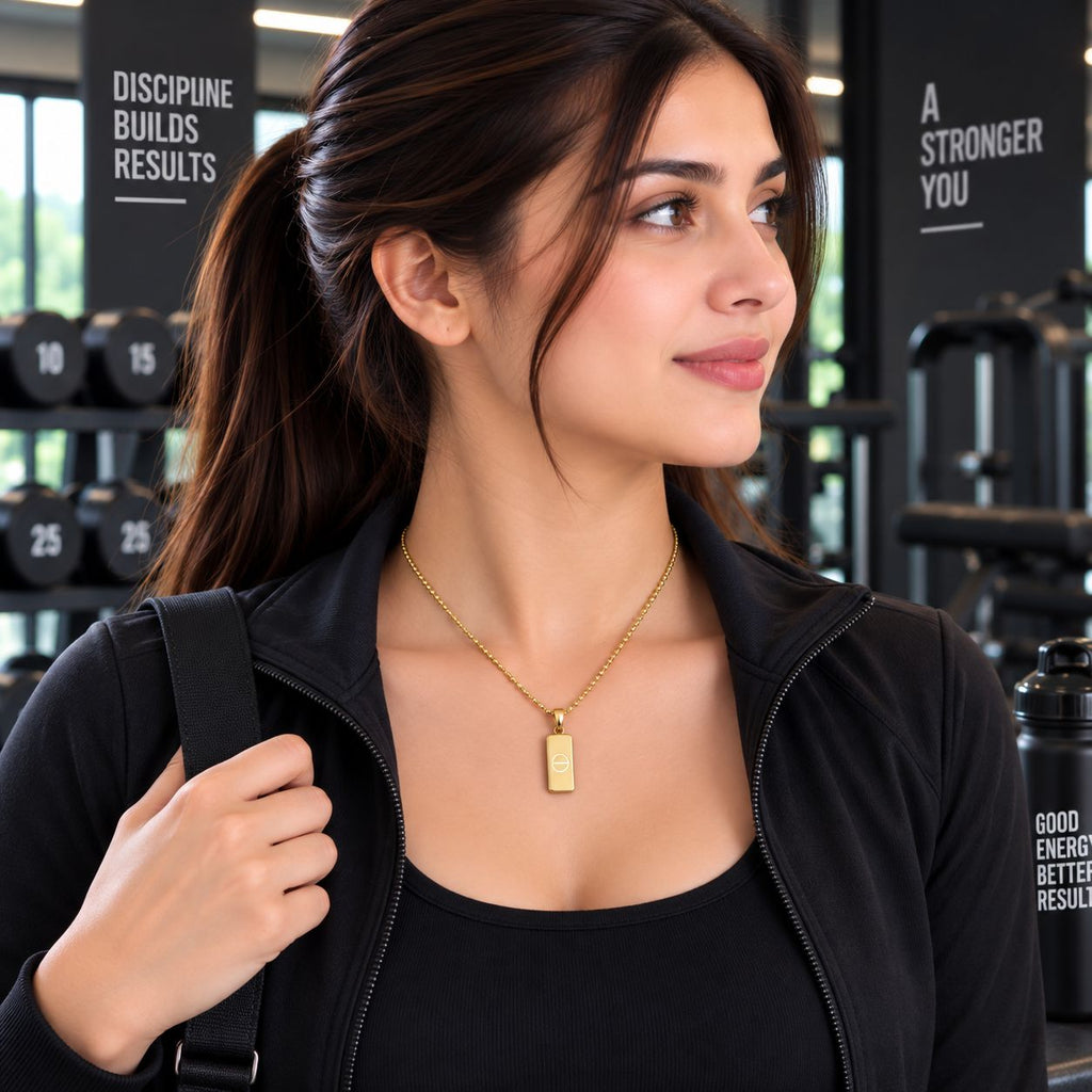 Gold Engraved Circle Dog Tag Pendant Necklace worn by model in black gym jacket at gym setting with dumbbell rack backdrop