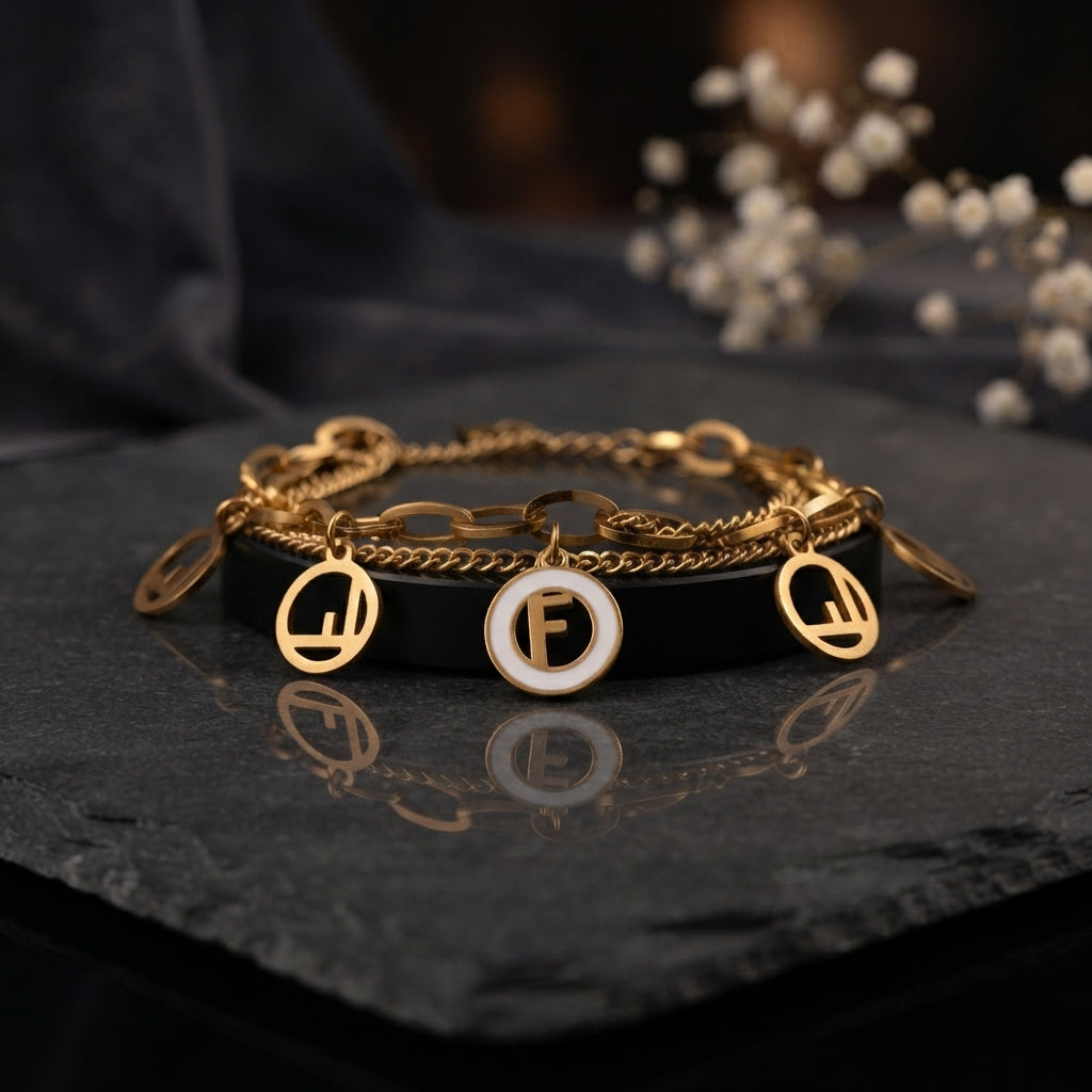 Gold F Logo Charm Double Layer Chain Bracelet in 18K gold-tone PVD stainless steel on dark slate surface with white gypsophila flowers and dark silk backdrop showing bold oval paperclip link chain layered with fine curb chain featuring 4 open-cut gold F logo circle disc charms and 1 white enamel F logo medallion centre charm with mirror reflection below