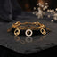 Gold F Logo Charm Double Layer Chain Bracelet in 18K gold-tone PVD stainless steel on dark slate surface with white gypsophila flowers and dark silk backdrop showing bold oval paperclip link chain layered with fine curb chain featuring 4 open-cut gold F logo circle disc charms and 1 white enamel F logo medallion centre charm with mirror reflection below