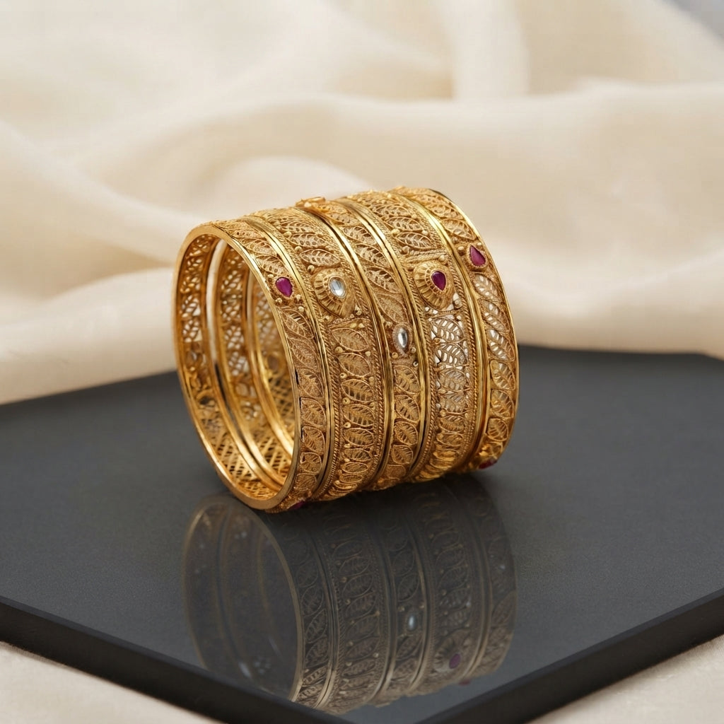 Gold Filigree Leaf Kundan Ruby Bangles Antique Set of 4 in 22K antique gold-tone plating on dark grey hexagonal mirror surface with ivory cream fabric backdrop showing four slim rigid bangles stacked together with full-coverage open gold filigree leaf paisley arabesque jali pattern and alternating clear polki kundan and ruby red cabochon accent stones in gold bezels with mirror reflection below