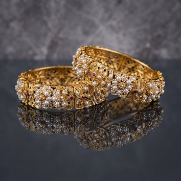 Gold Filigree Pearl Cluster Ruby Bridal Bangles Antique Pair in 22K antique gold-tone plating on dark grey mirror surface with marble backdrop showing two wide rigid bangles with dense floral pearl cluster surface featuring ruby red cabochon centres surrounded by oval rice pearls and seed pearl bead clusters set in ornate gold filigree arabesque leaf and scroll panels with mirror reflection below