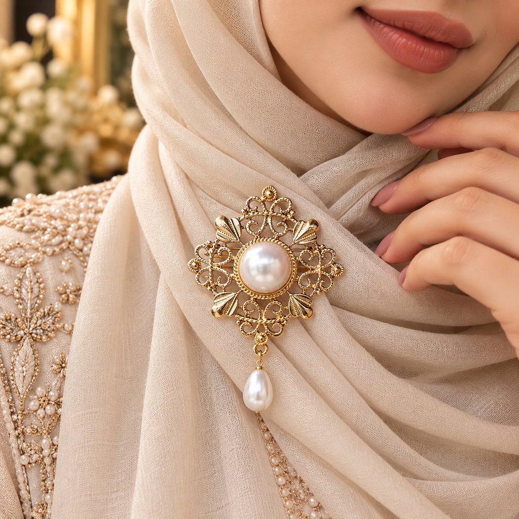 Gold Filigree Pearl Hijab Brooch Pin in antique 22K gold-tone worn on cream shimmer hijab in bridal editorial close-up showing ornate diamond-shaped arabesque filigree brooch with large round mabe pearl centre in rope-twist bezel four fan petal accents intricate scrollwork and baroque teardrop pearl drop pendant on gold embroidered bridal outfit backdrop