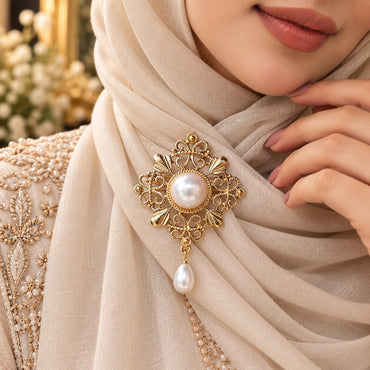 Gold Filigree Pearl Hijab Brooch Pin in antique 22K gold-tone worn on cream shimmer hijab in bridal editorial close-up showing ornate diamond-shaped arabesque filigree brooch with large round mabe pearl centre in rope-twist bezel four fan petal accents intricate scrollwork and baroque teardrop pearl drop pendant on gold embroidered bridal outfit backdrop