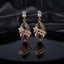 Gold Floral CZ Leaf Crystal Gem Drop Earrings in Ruby Red showing pair suspended on monofilament wire against dark backdrop with baguette CZ stud top large faceted diamond-shape ruby red crystal drop with gold daisy flower and marquise CZ leaf sprig overlay and mirror reflection below