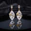 Gold Floral CZ Leaf Crystal Gem Drop Earrings in Emerald Green showing pair suspended on monofilament wire against dark burgundy velvet backdrop with baguette CZ stud top large faceted diamond-shape emerald green crystal drop with gold daisy flower and marquise CZ leaf sprig overlay and mirror reflection below