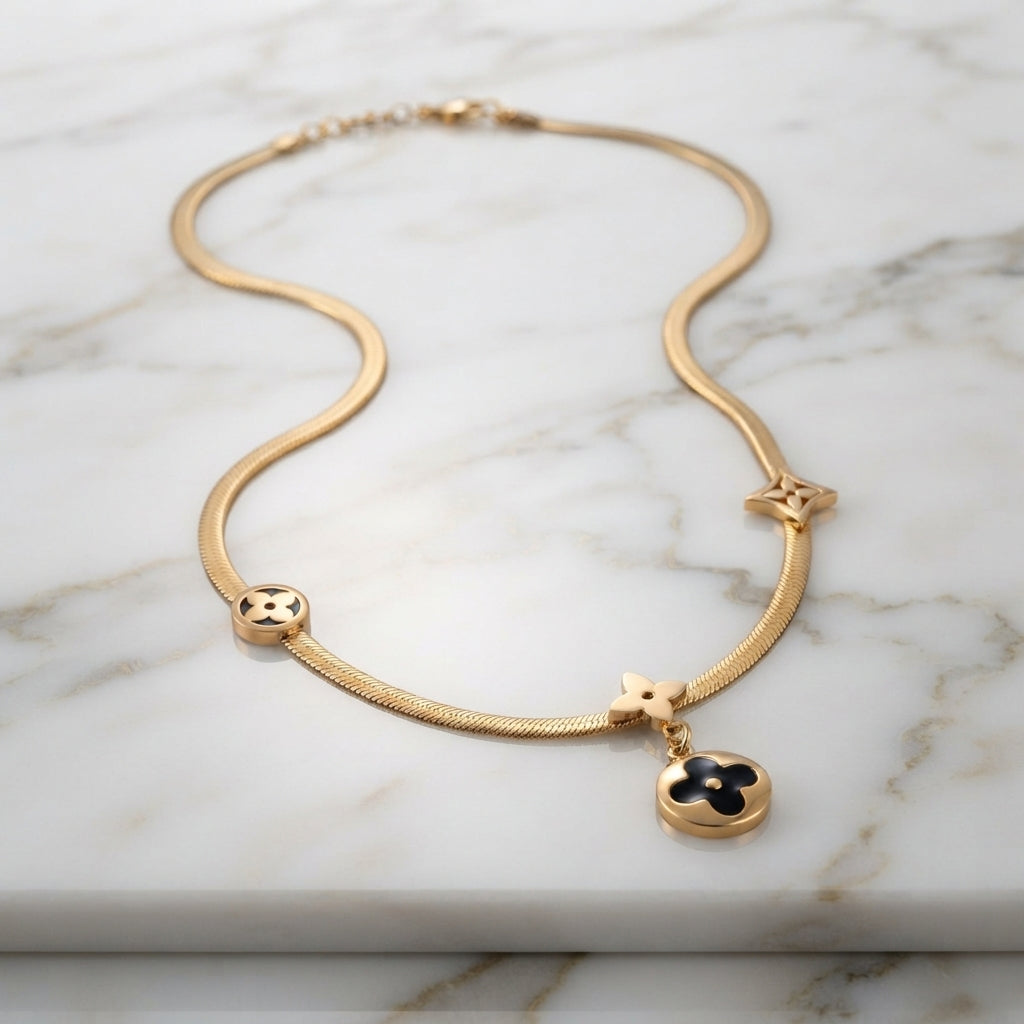Gold Four-Leaf Clover Monogram Herringbone Chain Pendant Necklace draped on dark grey satin silk fabric backdrop with dark mirror surface showing 18K yellow gold-tone flat herringbone snake chain with three four-leaf clover monogram flower charms including hanging round black enamel disc drop pendant with mirror reflection below
