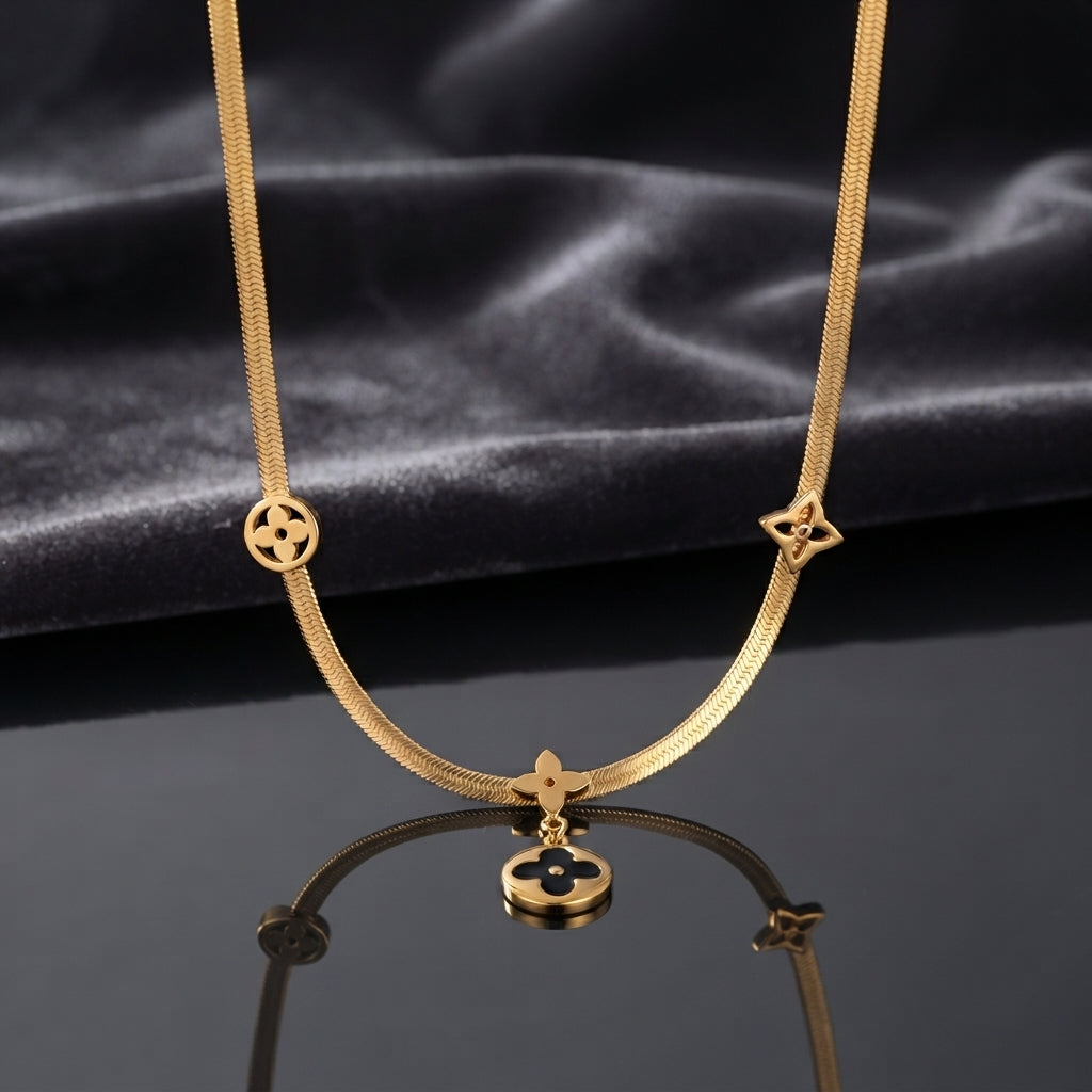 Gold Four-Leaf Clover Monogram Herringbone Chain Pendant Necklace laid flat on white Carrara marble surface in warm natural light showing 18K yellow gold-tone flat herringbone snake chain with round black enamel disc charm quatrefoil star outline charm and hanging round black enamel disc drop pendant with four-petal clover motifs throughout
