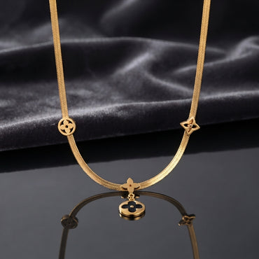 Gold Four-Leaf Clover Monogram Herringbone Chain Pendant Necklace laid flat on white Carrara marble surface in warm natural light showing 18K yellow gold-tone flat herringbone snake chain with round black enamel disc charm quatrefoil star outline charm and hanging round black enamel disc drop pendant with four-petal clover motifs throughout